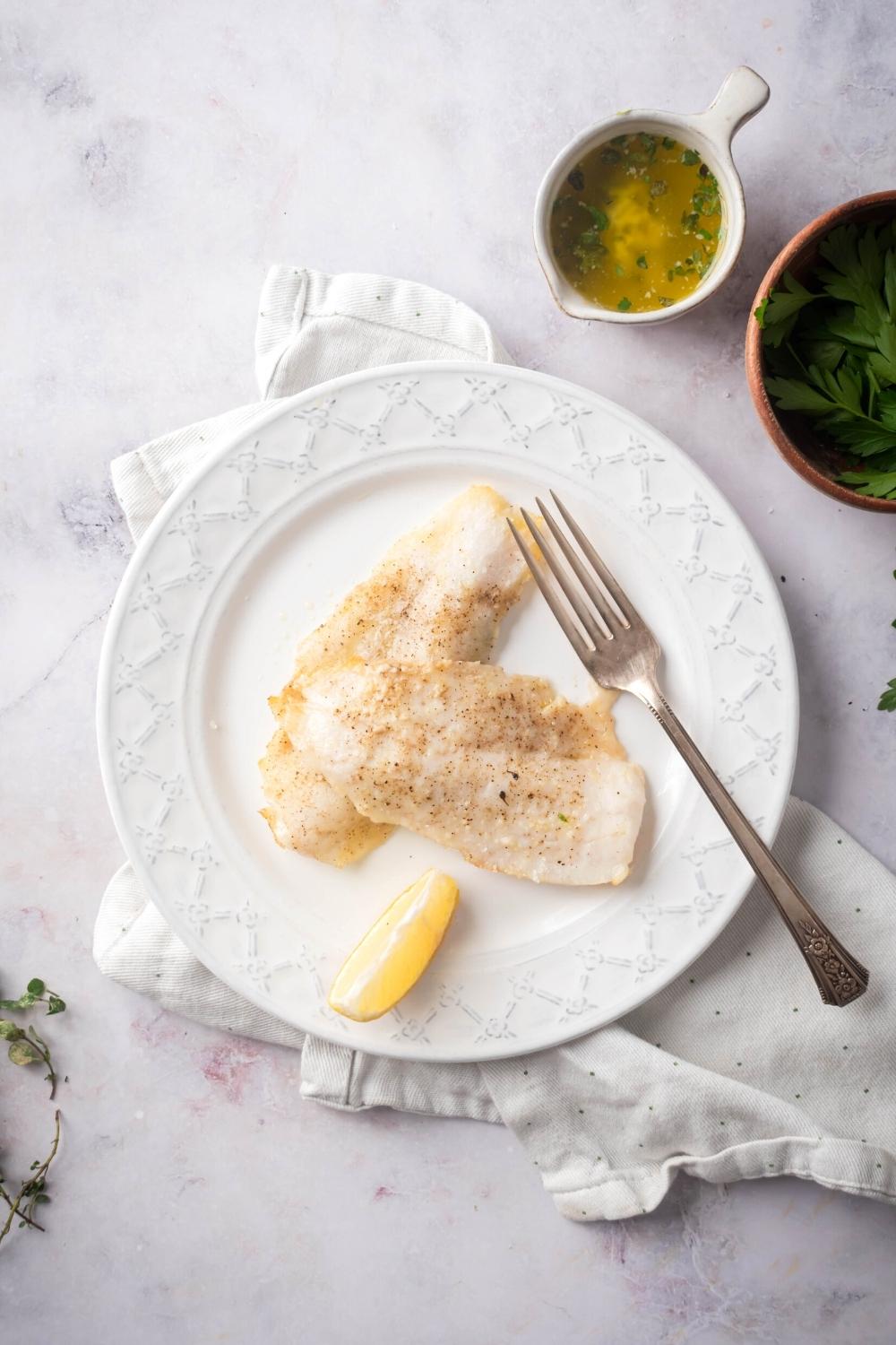 Two mahi mahi fillets, a fork, and a lemon wedge on top of a white plate on a white tablecloth on a grey counter. Behind it is a small white cup filled with garlic butter.
