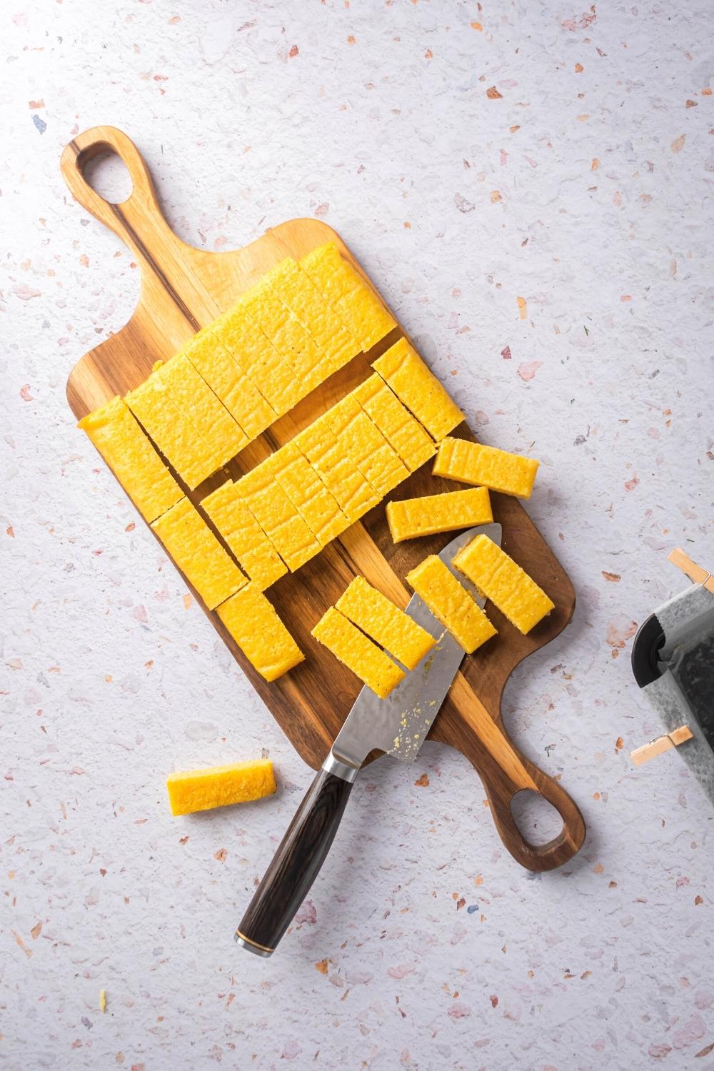 Slices of polenta on top of the wooden cutting board with a knife set at the front of the cutting board covered with a few polenta sticks.