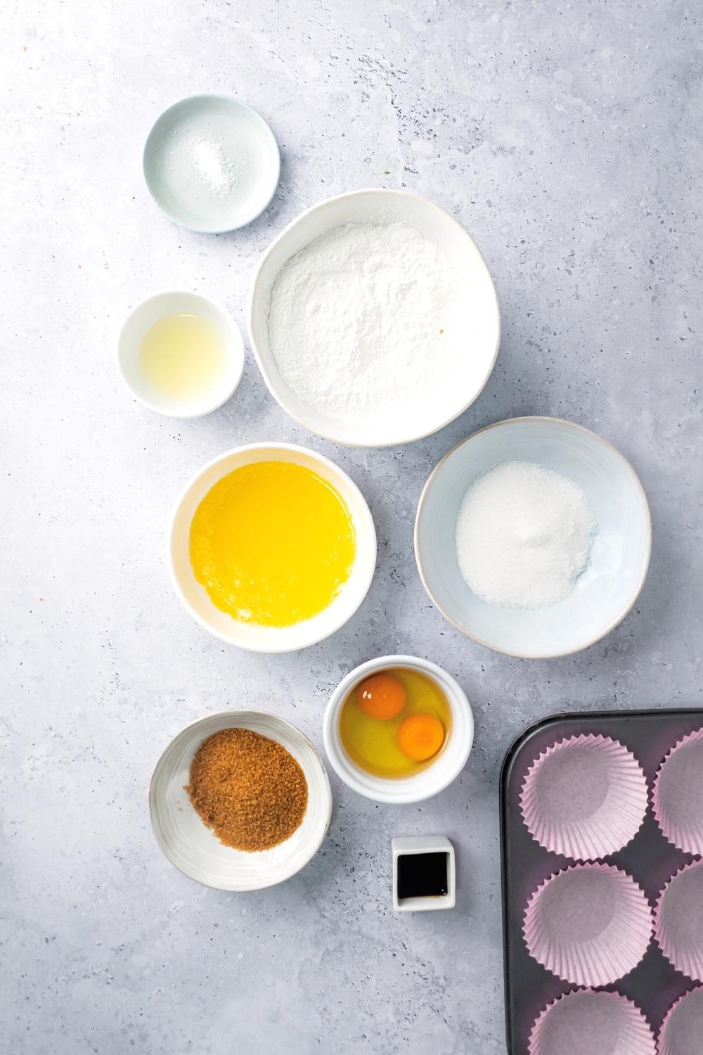 A bowl of brown sugar, a bowl of two eggs, a bowl of sugar, a bowl of melted butter, a bowl of flour, and a bowl of salt all on a white counter.