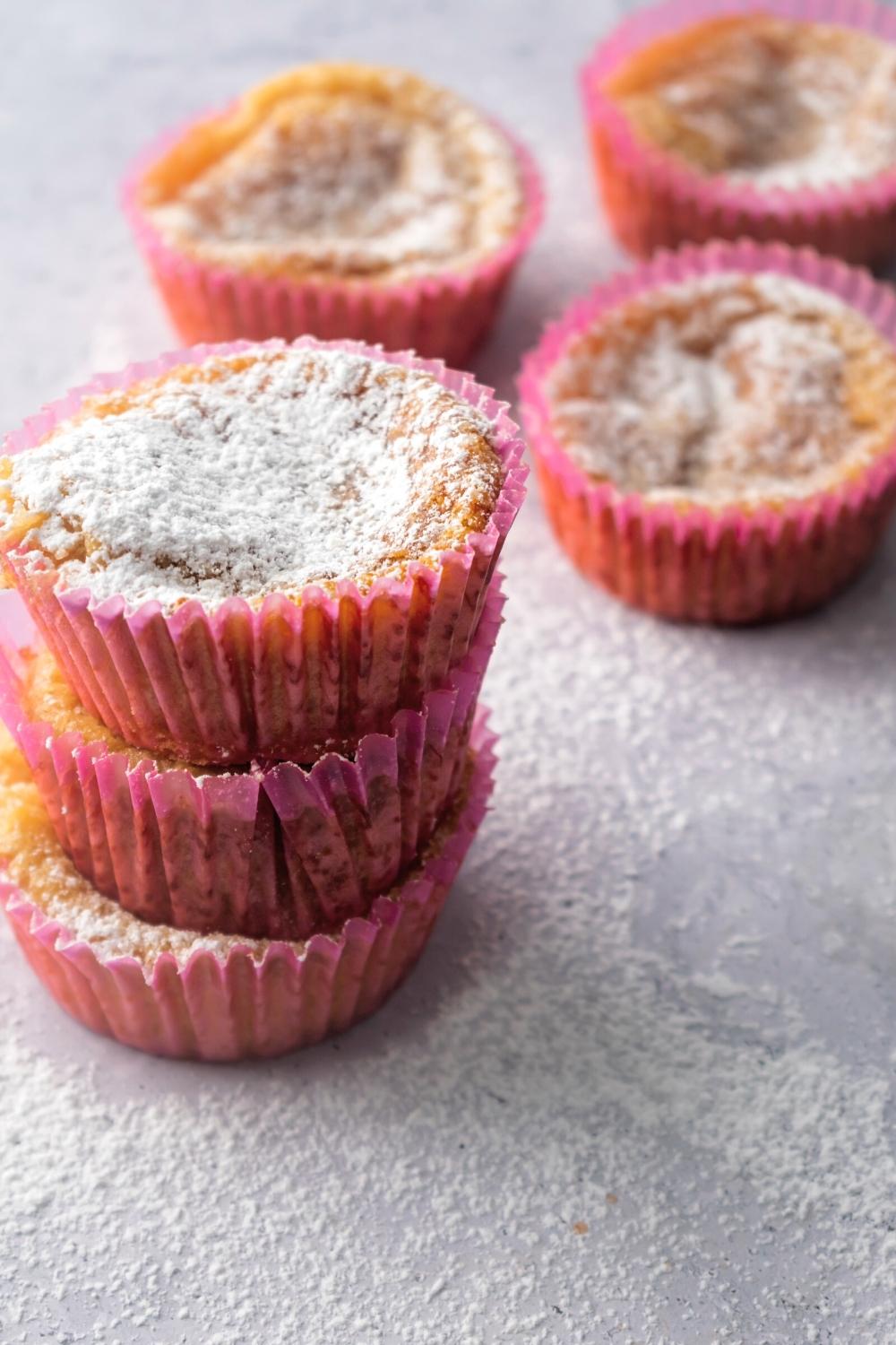 A stack of three mochi muffins with powdered sugar on the top one. Behind it is three more mochi muffins on a grey counter.