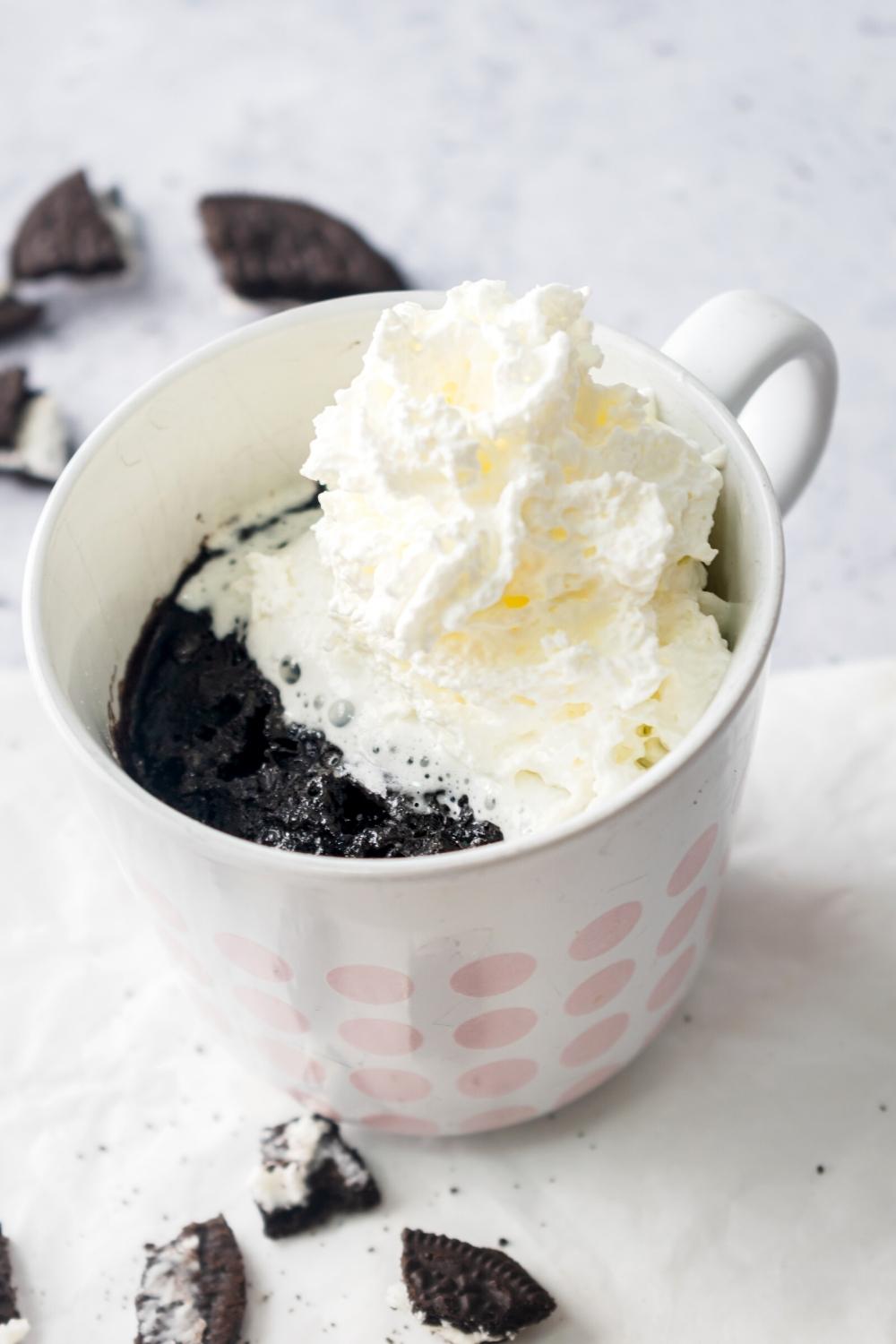 A Oreo mug cake in a white bag with a dollop of whipped cream and half of it. There are some pieces of Oreo cookie around the mug.