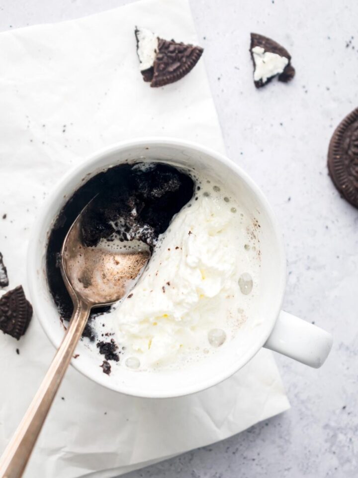Whipped cream and a spoon on top of an Oreo mug cake in a white bag that is on a white napkin on the white counter.
