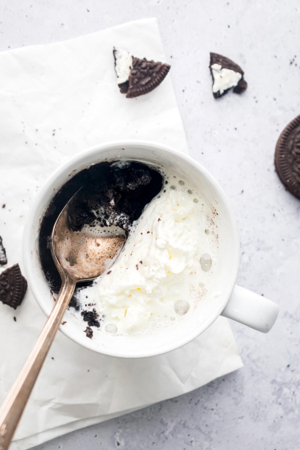 Whipped cream and a spoon on top of an Oreo mug cake in a white bag that is on a white napkin on the white counter.