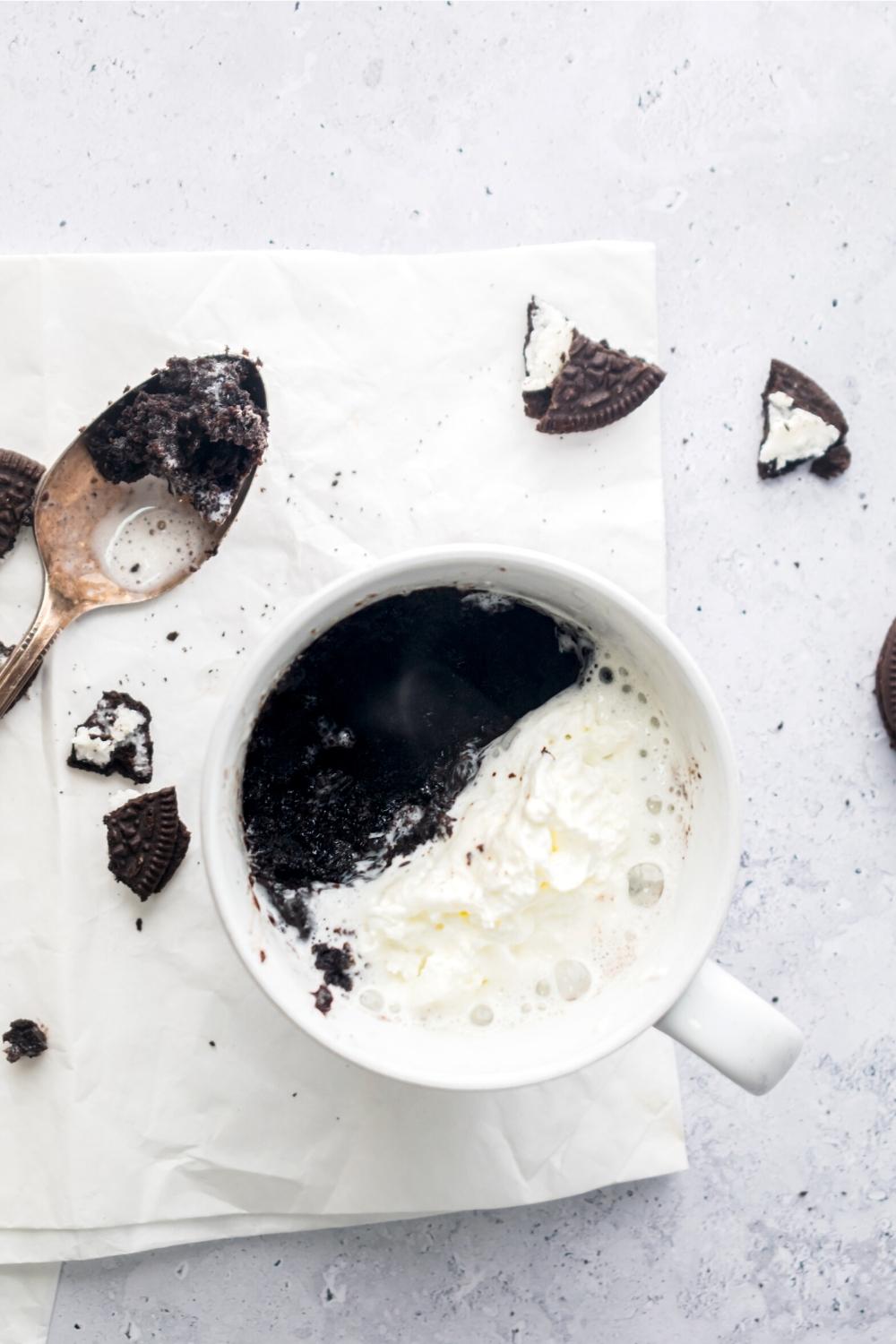 Half whipped cream and a half Oreo mug cake in a white bag on a napkin or a white counter. There's a spoon covered and Oreo mug cake and some pieces of Oreo around the mug.