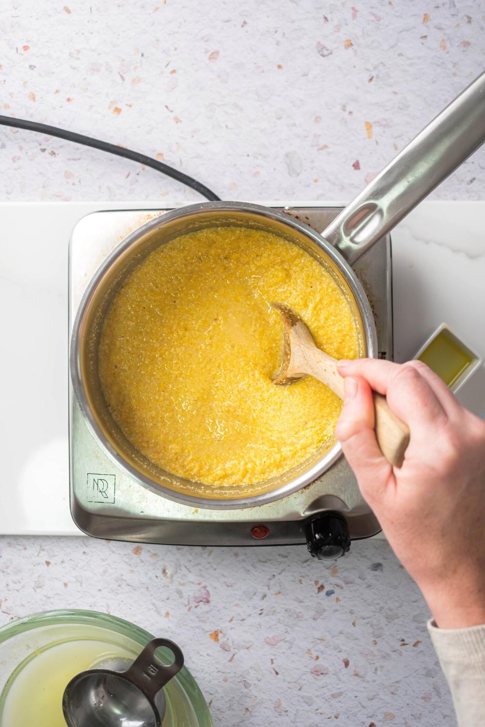 A hand holding a wooden spoon that is submerged in a pot filled with creamy polenta. The pot is on a burner that is set on a white counter.
