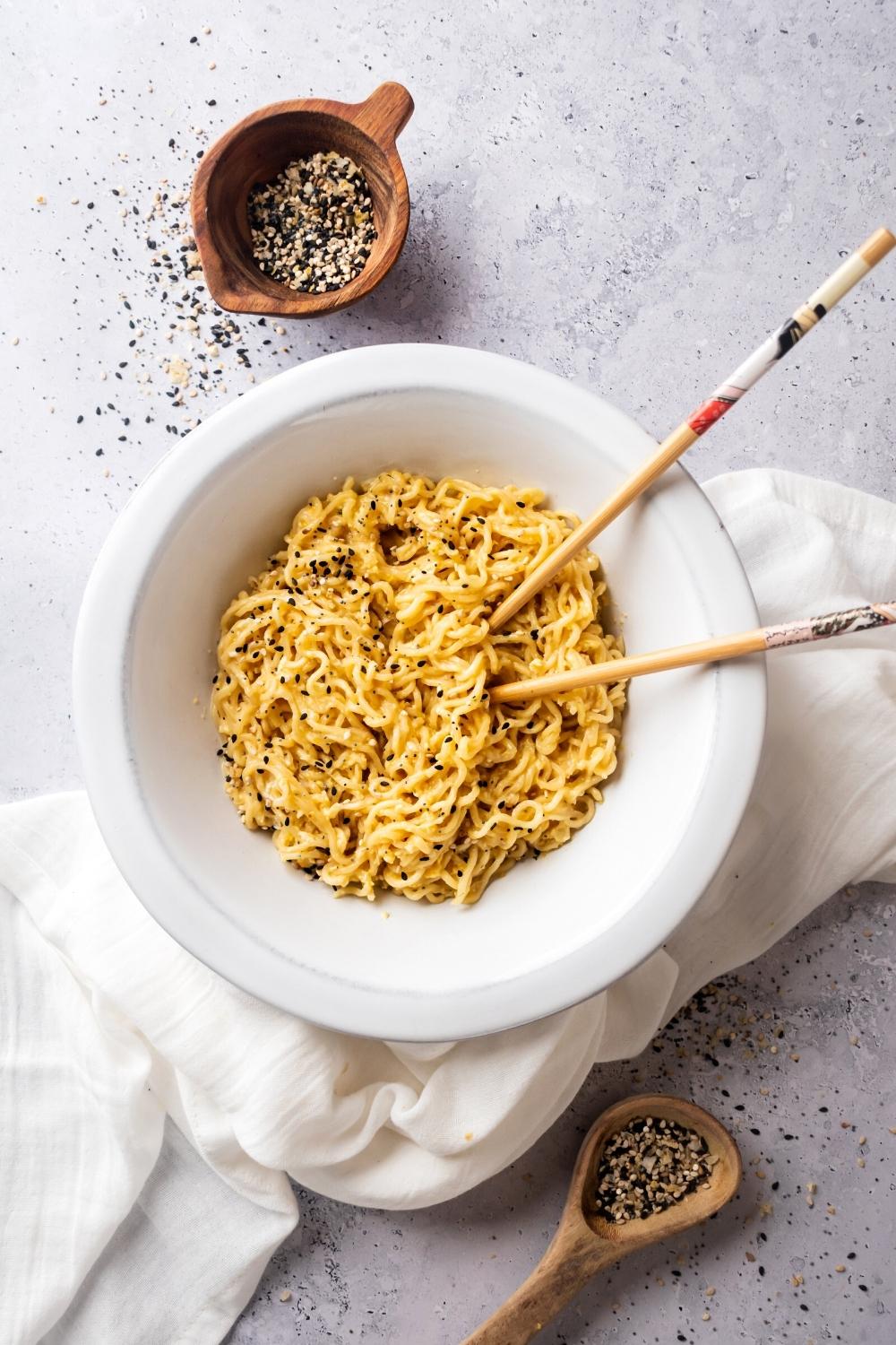 Ramen noodles in a white bowl and part of a white tablecloth and a gray counter. Part of to Chopstix or submerged in the noodles and in front of and behind the white bowl or two wooden bowls filled with everything bagel seasoning.