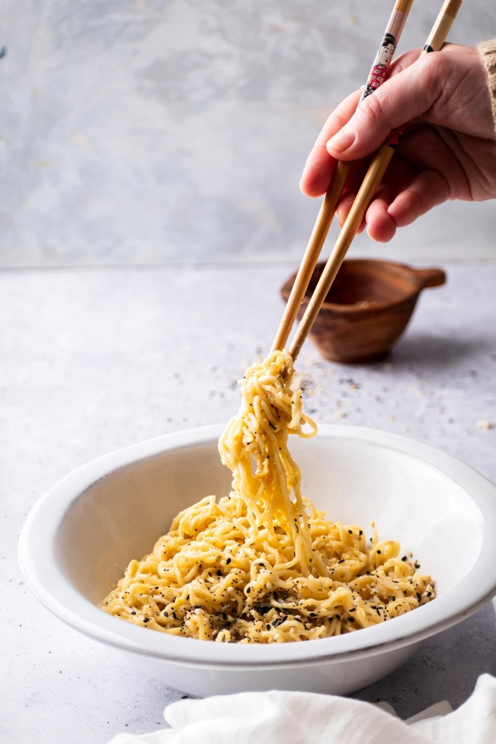 A hand holding a pair of chopsticks with Ramen noodles pinched between the two. The noodles are being held over on white bowl filled with Ramen noodles.