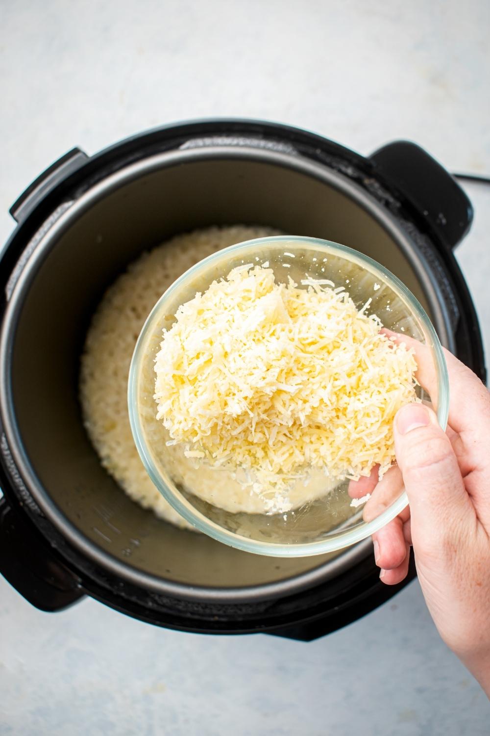 A hand holding a bowl of Parmesan cheese over an instant pot filled with risotto.