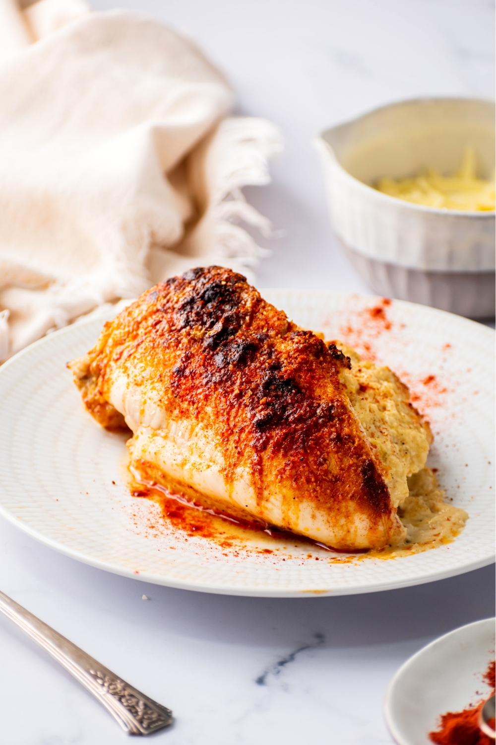 A stuffed chicken breast on a white plate. Behind it is part of a white bowl filled with cheddar cheese.