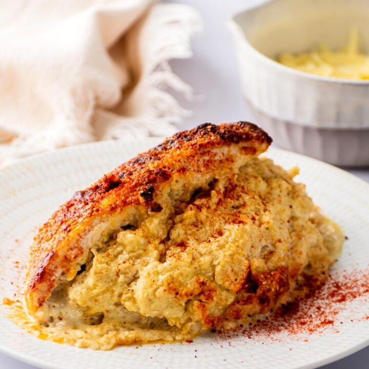 A chicken breast sliced in half stuffed with filling. The chicken is on a white plate and behind it is part of a white bowl with cheddar cheese in it.