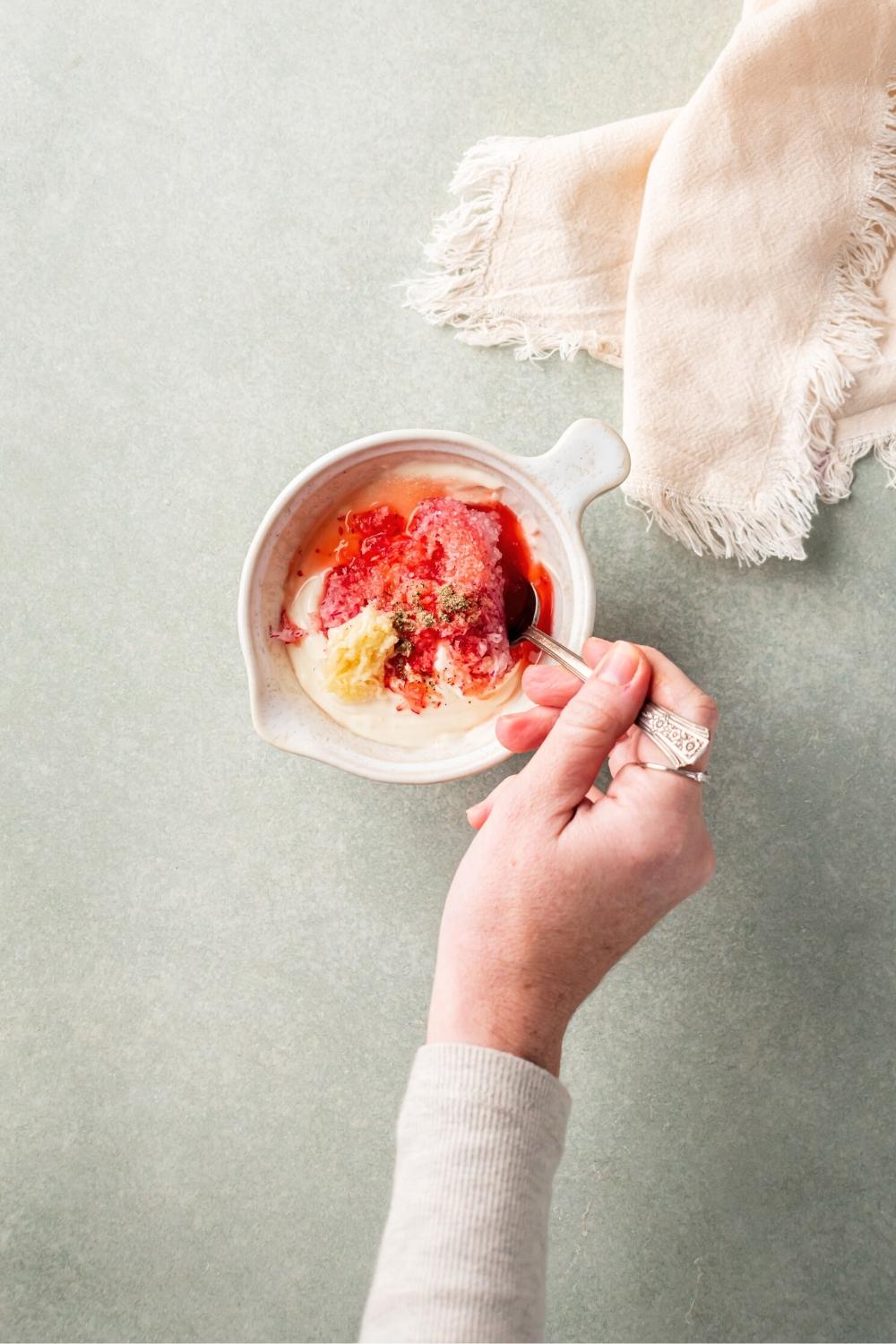 A hand holding a spoon mixing tiger sauce ingredients in a white bowl on a gray counter