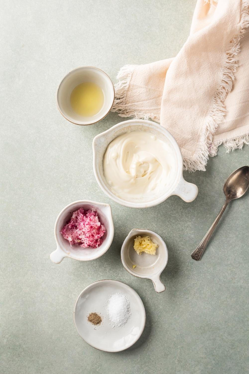 A white plate with some salt and pepper on it, a bowl of garlic, a bowl of horseradish, a big bowl of mayo, and a small bowl of oil on a gray counter.