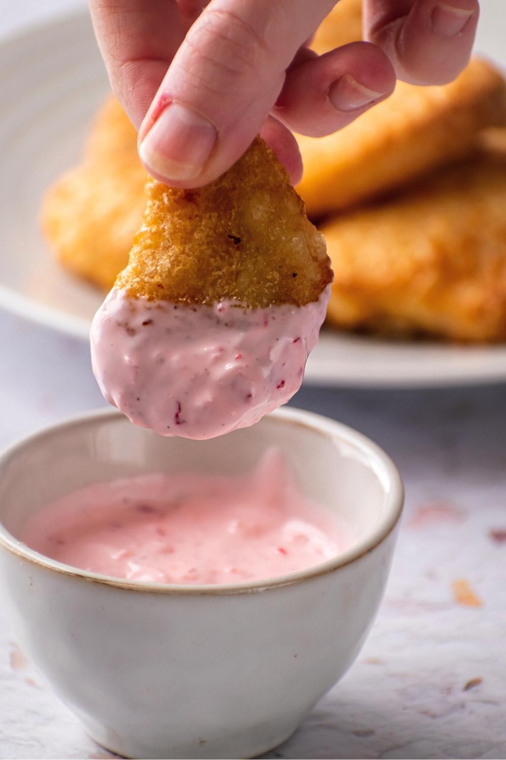 Fingertips holding a chicken nugget that has tiger sauce on it over a white bowl filled with the tiger sauce. In the background is part of a white plate with chicken nuggets on it.