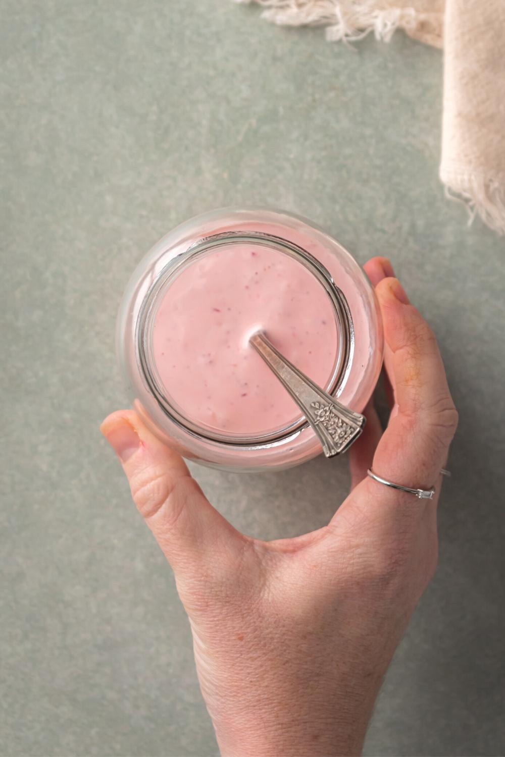 A hand holding a glass jar that is filled with tiger sauce with the utensil submerged in it. The glass jars on top of the gray counter.