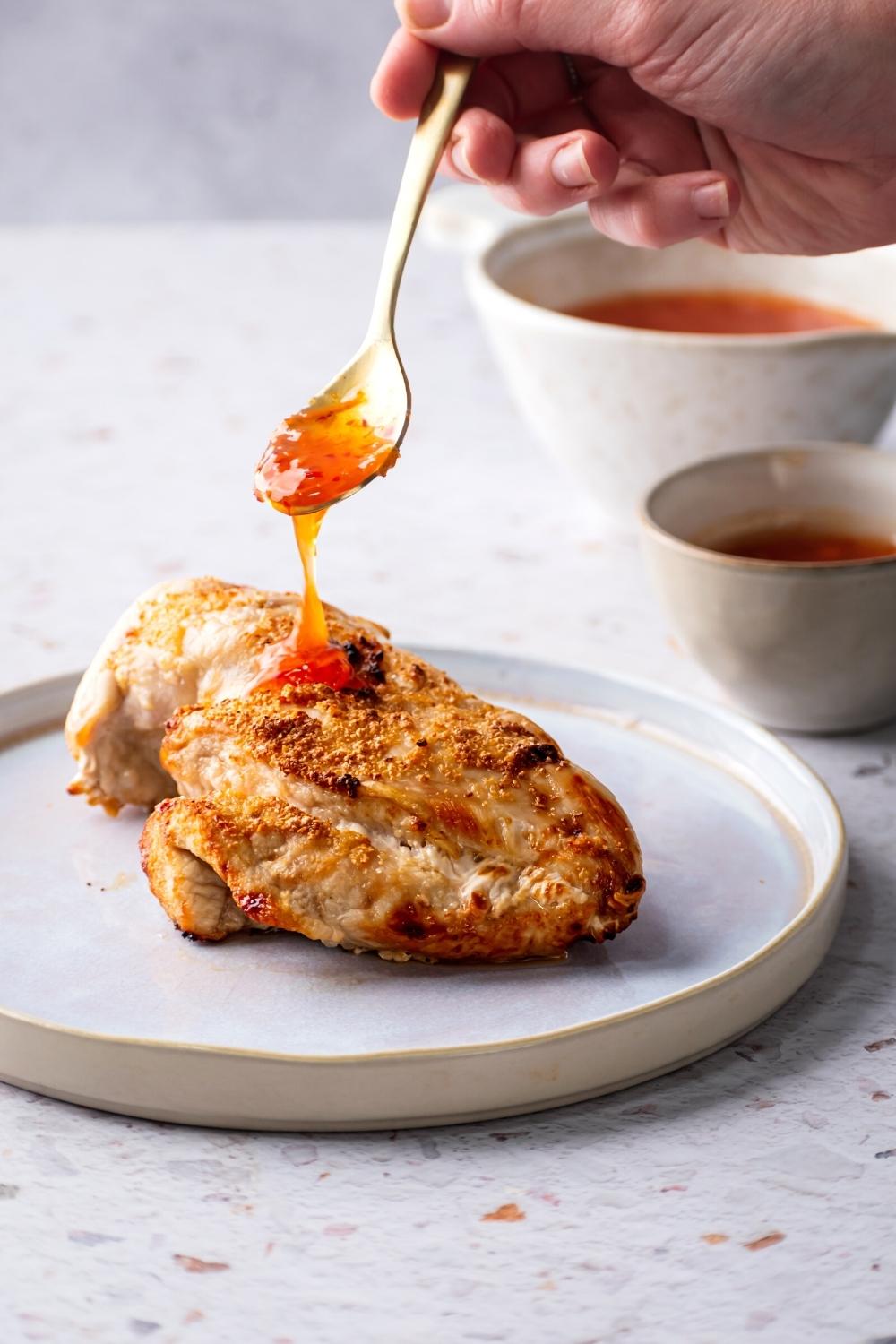 A hand drizzling sweet chili sauce on a piece of chicken breast on a white plate.