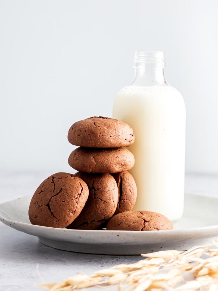 Six Nutella cookies that are stacked on top of one another leaning against a glass jar of milk all on a white plate on a white counter.