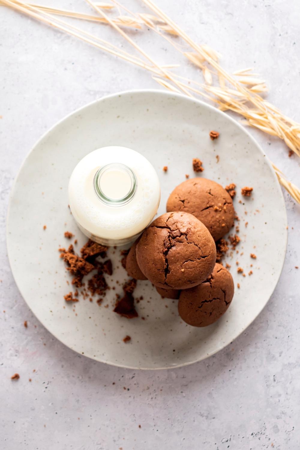 A couple of Nutella cookies stacked on top of one another on top of the plate that is on a white counter. Next to the cookies on the plate is a glass jar of milk.