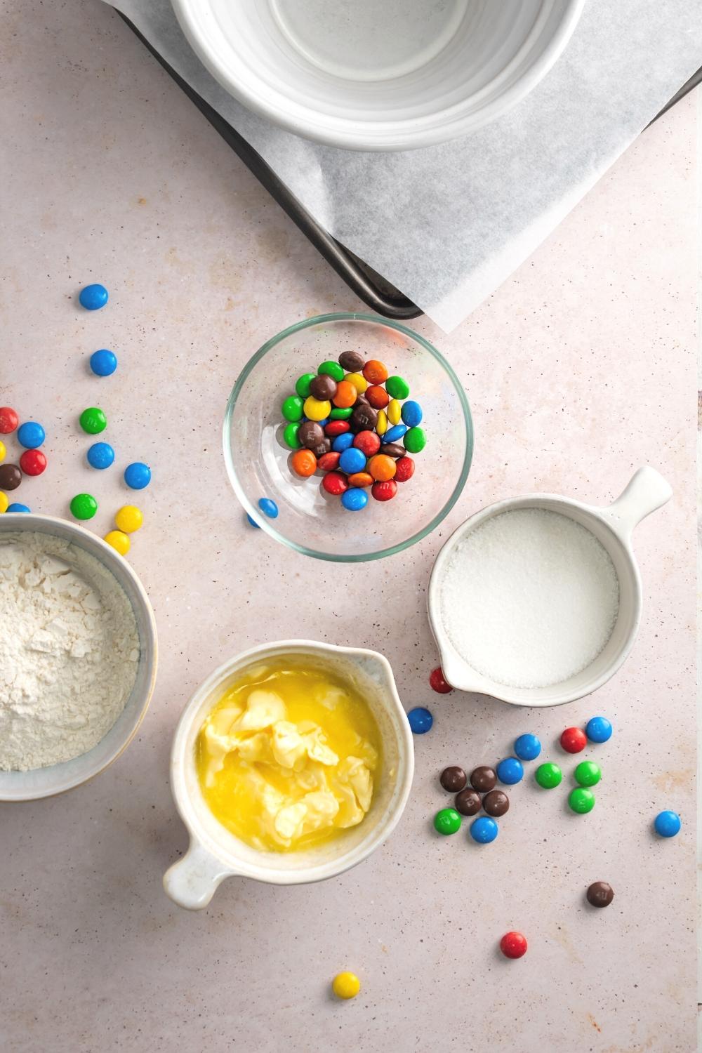 A bowl of melted butter, a bowl of flour, bowl of sugar, and a bowl of M&Ms all on a pink counter.