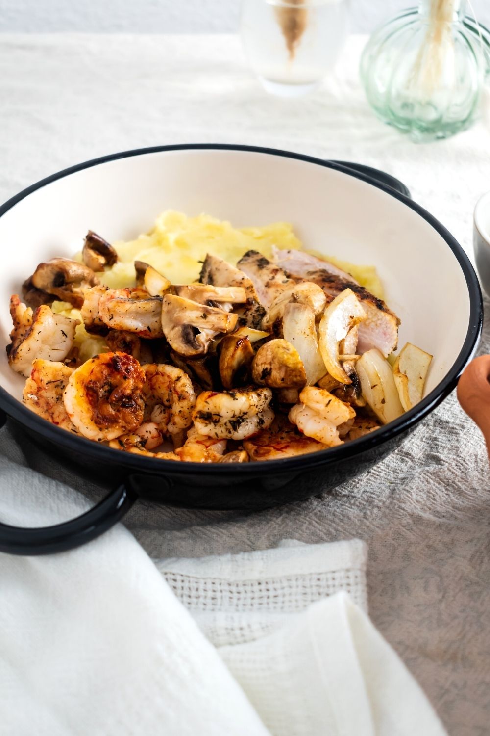 A bowl filled with grilled chicken, shrimp, mushrooms, and potato purée on a gray tablecloth.