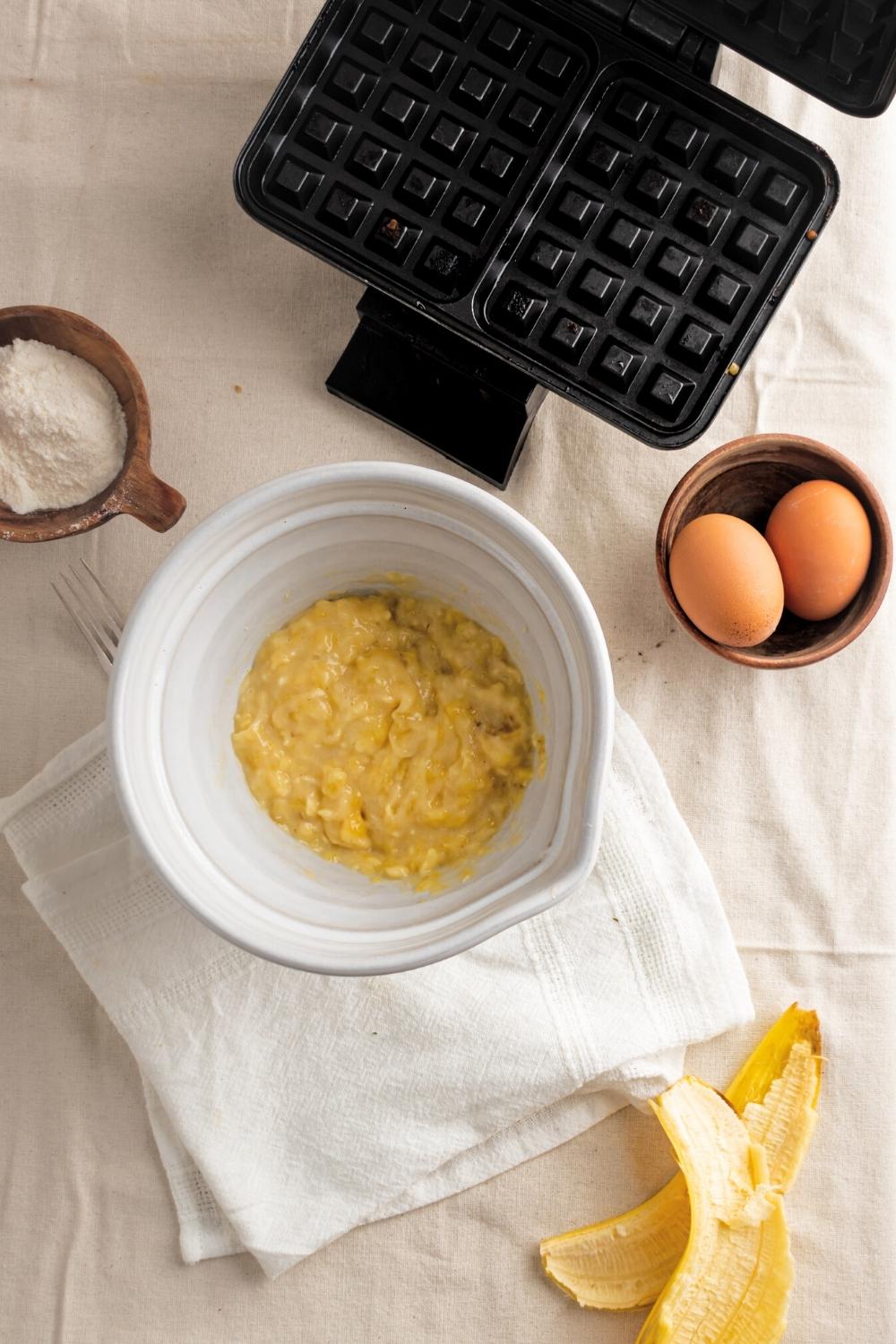 Mashed bananas in a bowl, two eggs in a small bowl, a small bowl of coconut flour, and part of a waffle iron on a white tablecloth.
