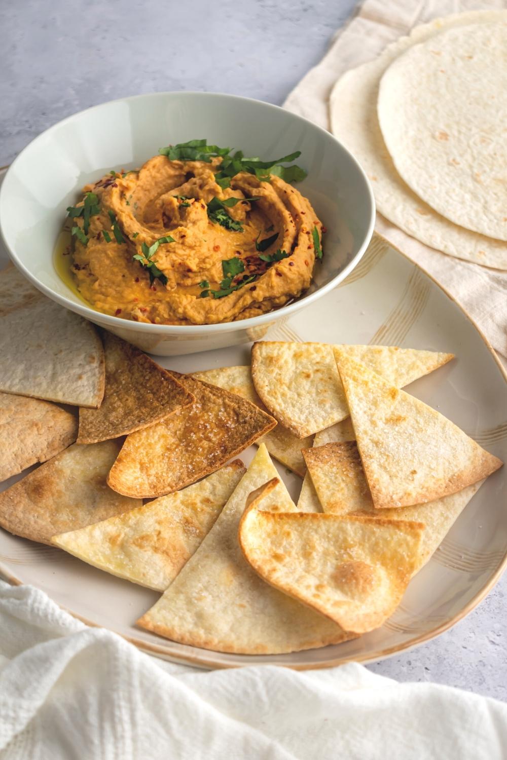 A bunch of air fryer tortilla chips overlapping one another on an oval shaped plate. On the plate behind the chips is a white bowl filled with hummus.