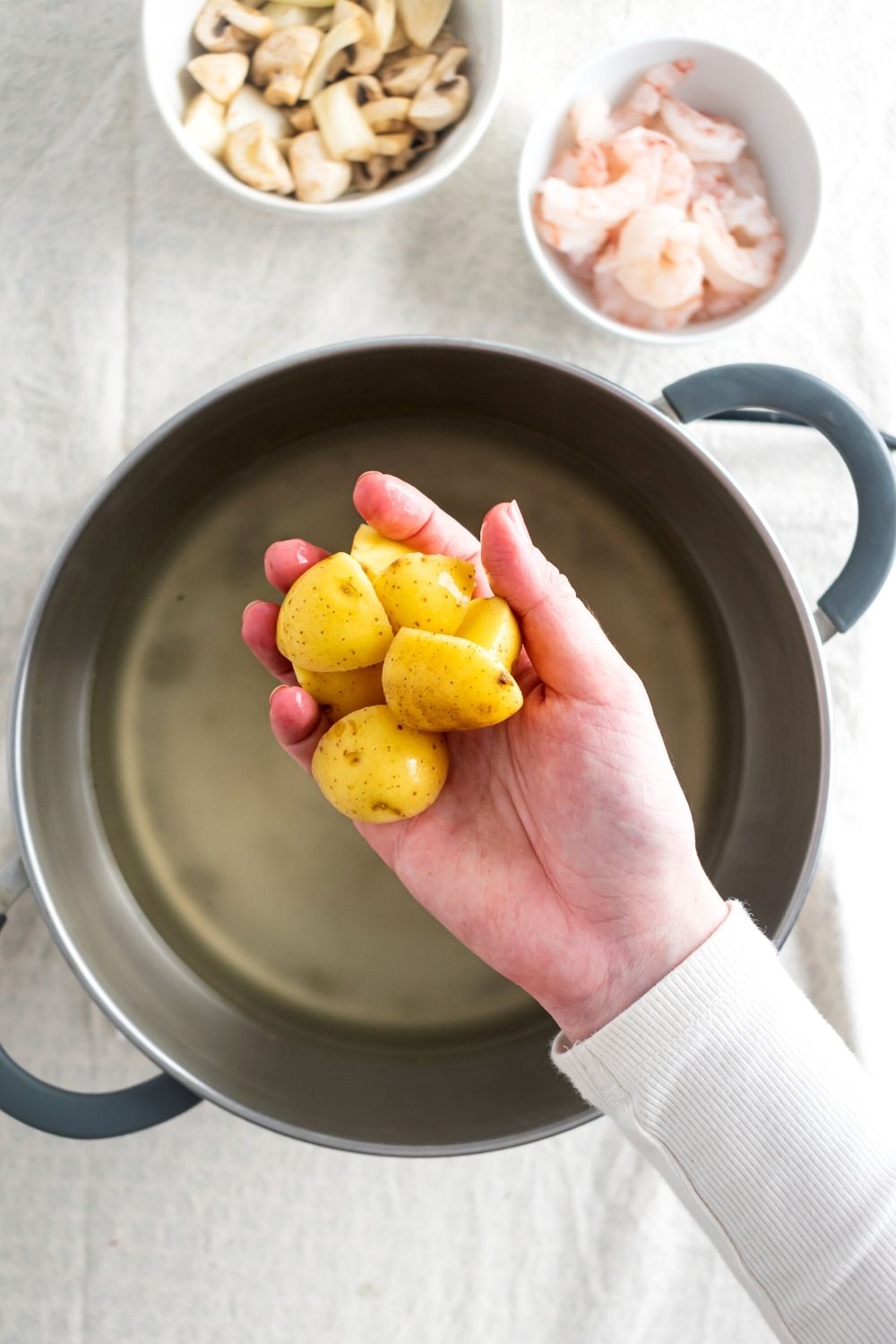 Hey hand holding small golden potatoes over a pot of water. Behind a pot of water is a white bowl of rice shrimp and part of a white bowl of mushrooms.
