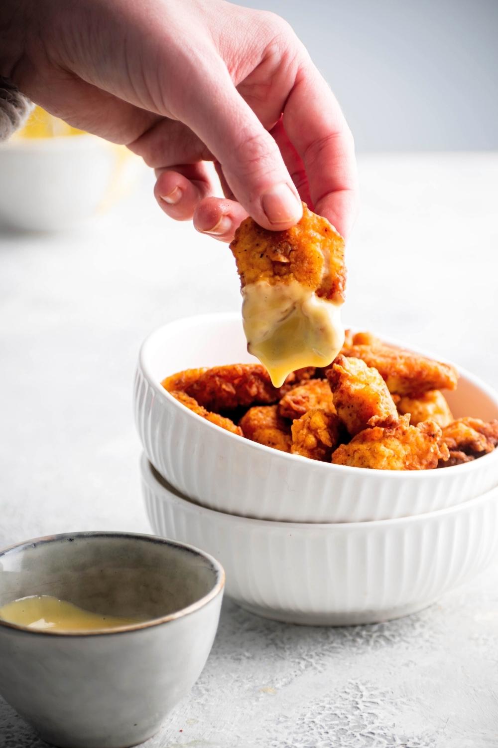 A hand holding a Chick-fil-A nugget with Chick-fil-A sauce on it over a white bowl that is filled with the Chick-fil-A nuggets.