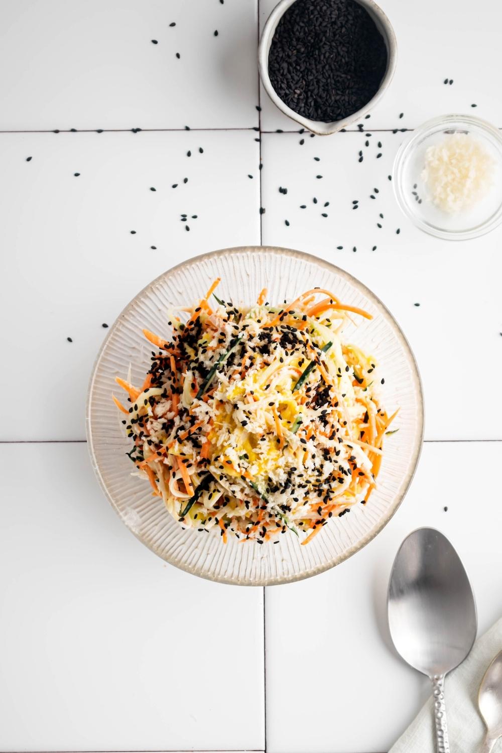 Kani salad in a glass bowl on a white tile counter. Beind it is a white cup of black sesame seeds.