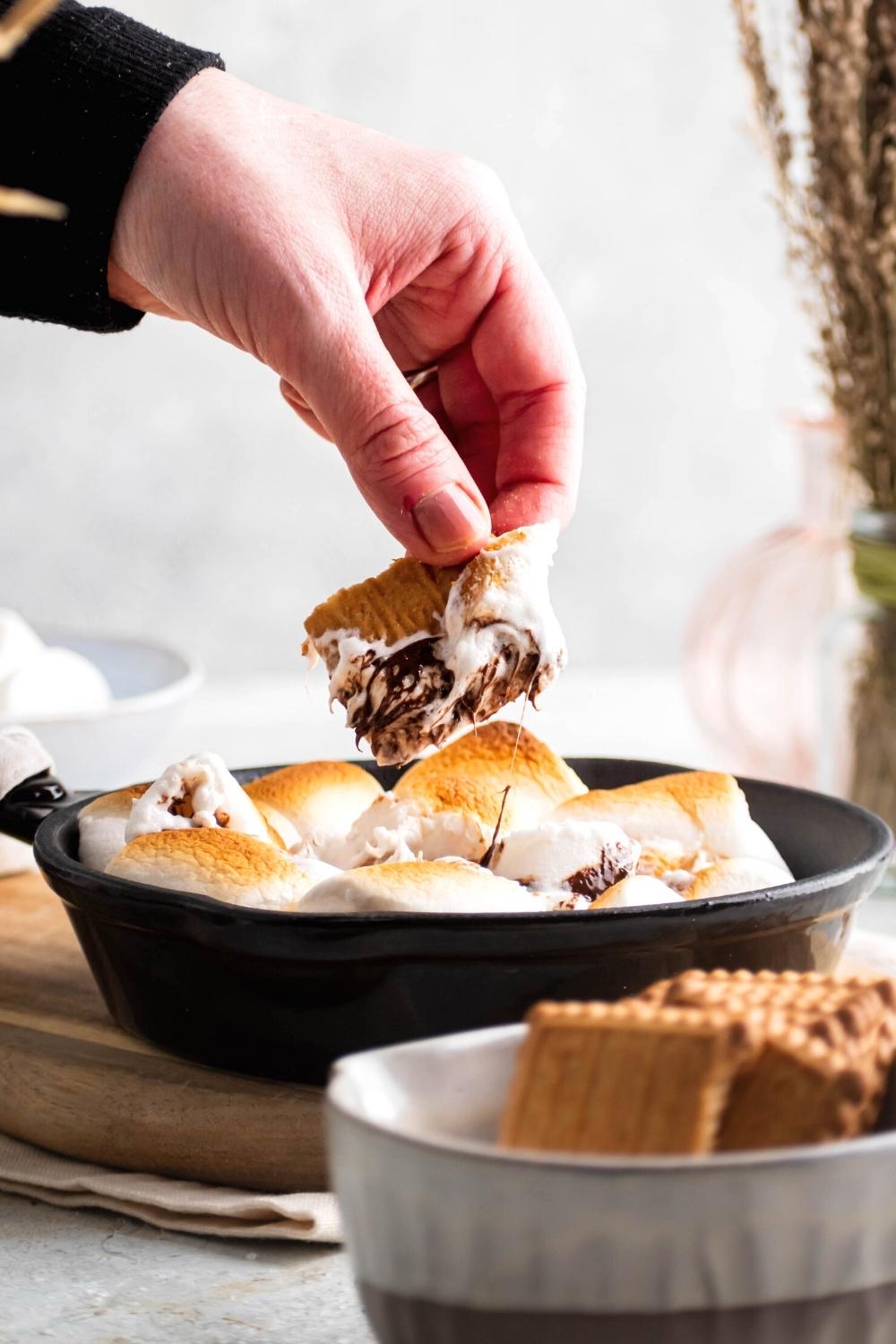 A hand holding a graham cracker that has some marshmallow and chocolate on it. The hand is holding it above toasted marshmallows and melted chocolate in a cast iron skillet.