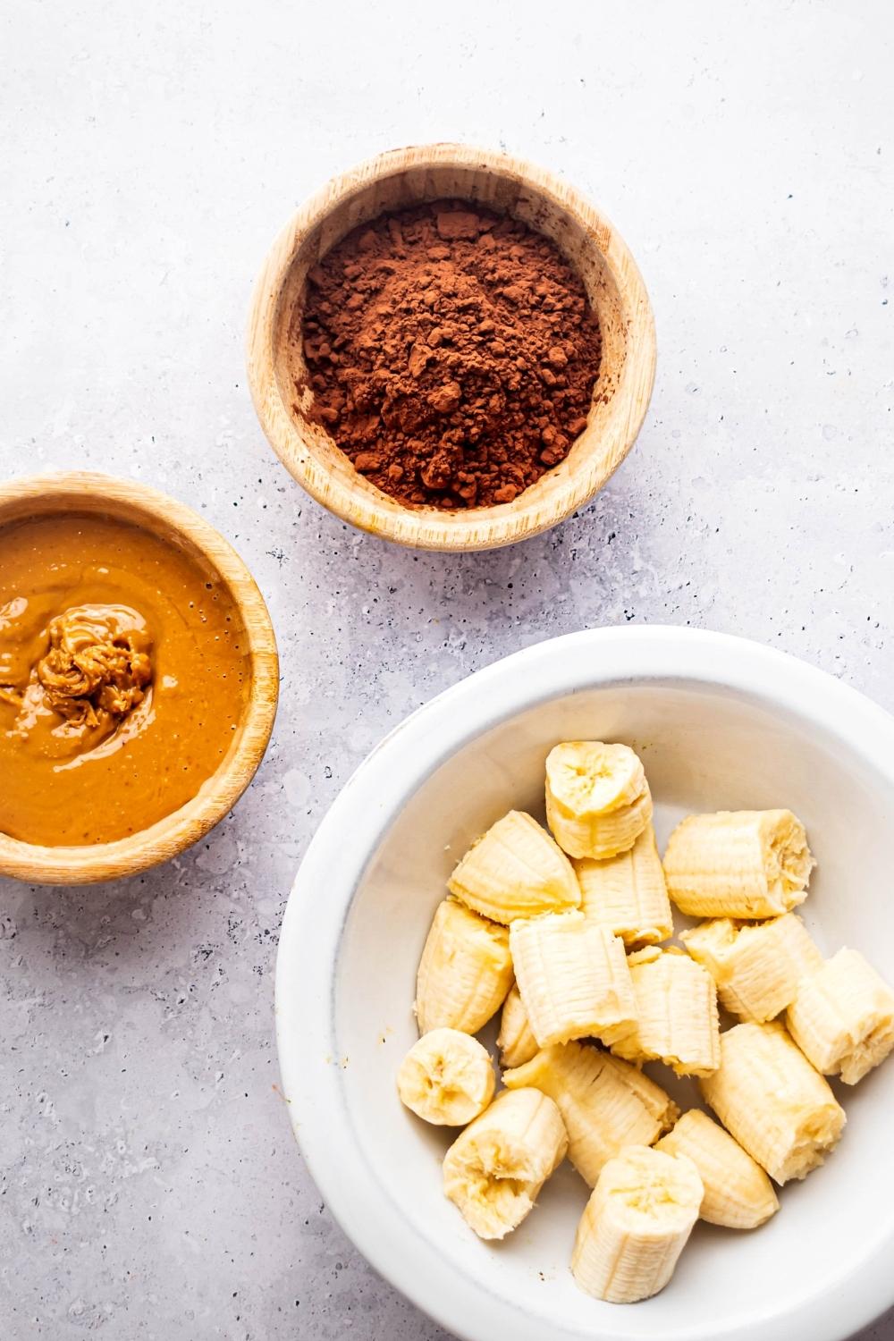 Pieces of banana and white bowl, creamy peanut butter in part of a bowl, and cocoa powder in a bowl all on a white counter.