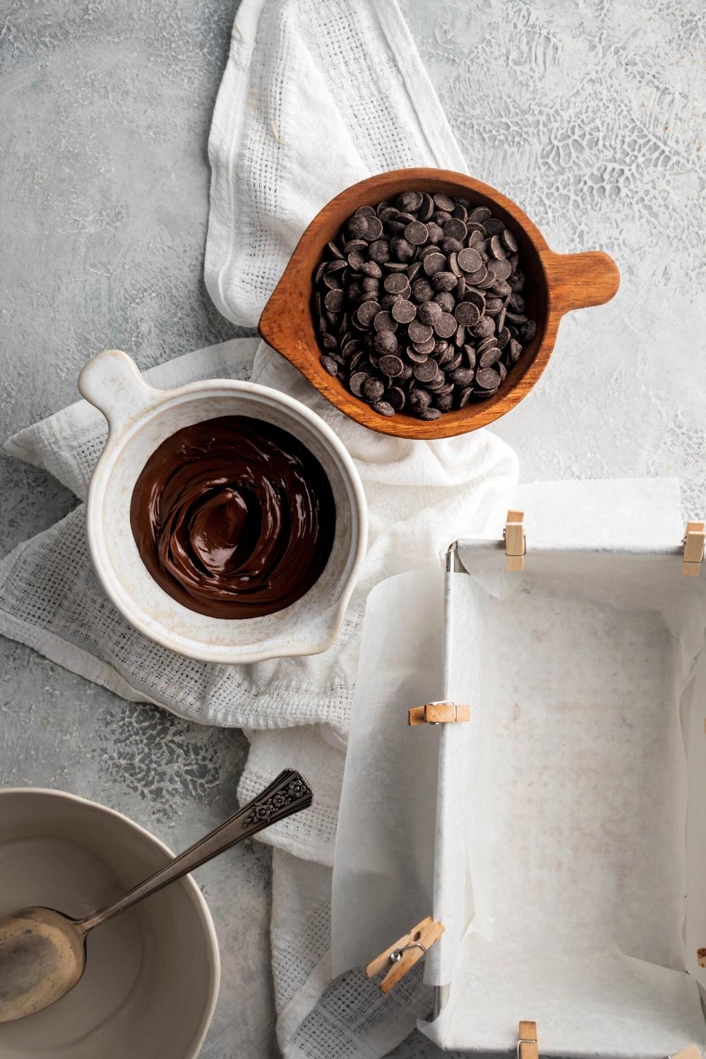 A wooden bowl of chocolate chips and a white bowl of Nutella on a white cloth on top of a grey counter.