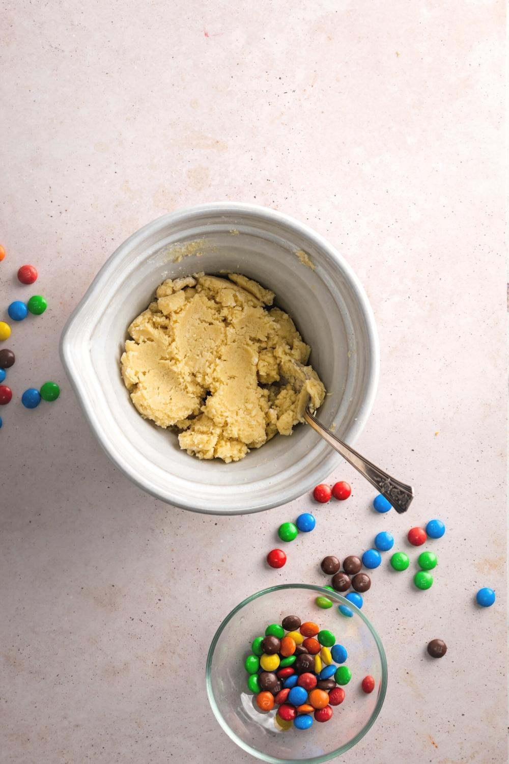 Sugar cookie dough in a white bowl with a spoon submerged in it. Behind it is a glass bowl with M&Ms and some M&Ms are on the table surround in the bowls..