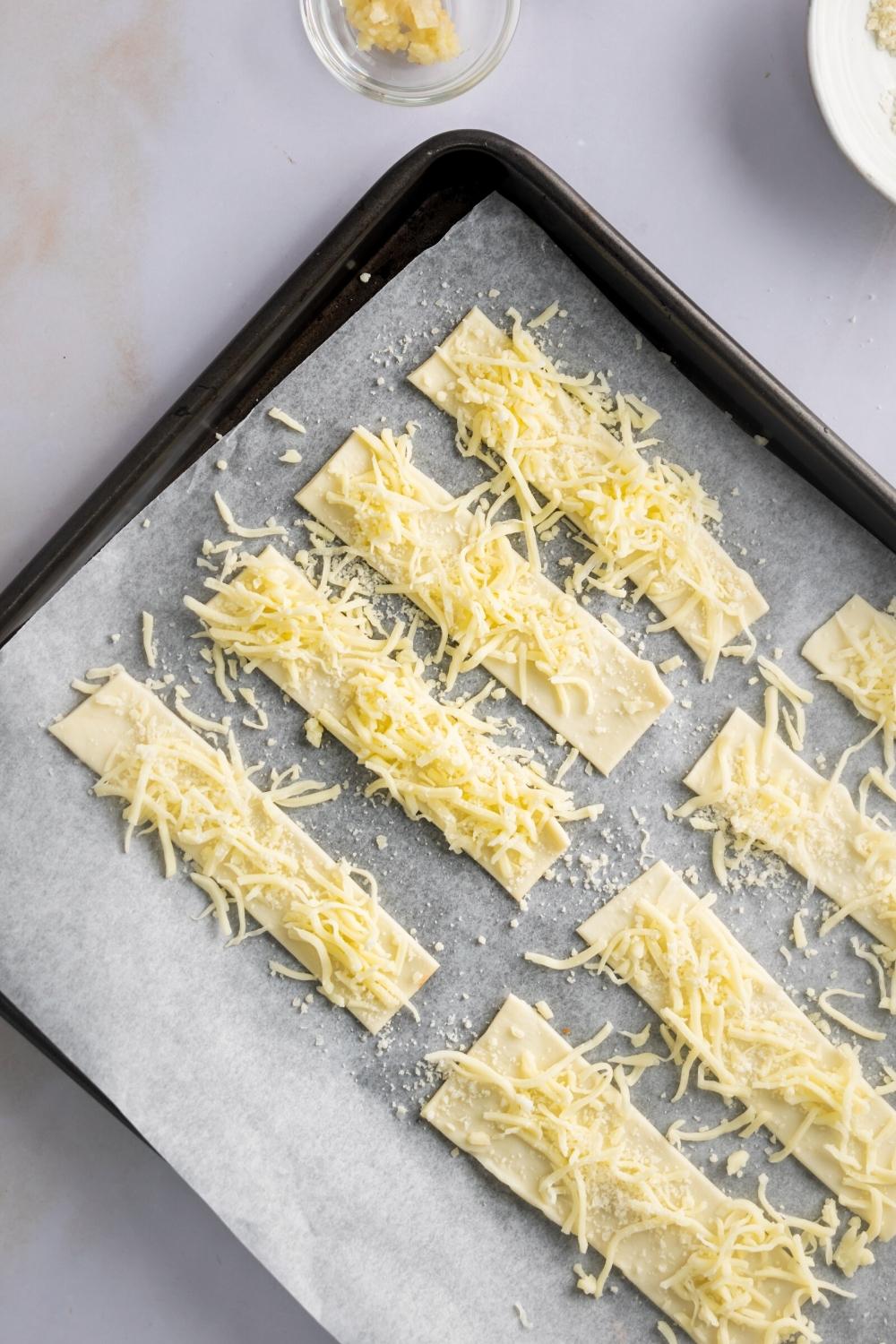 Shredded cheese on top of four dough sticks on a piece of parchment paper on a baking tray.