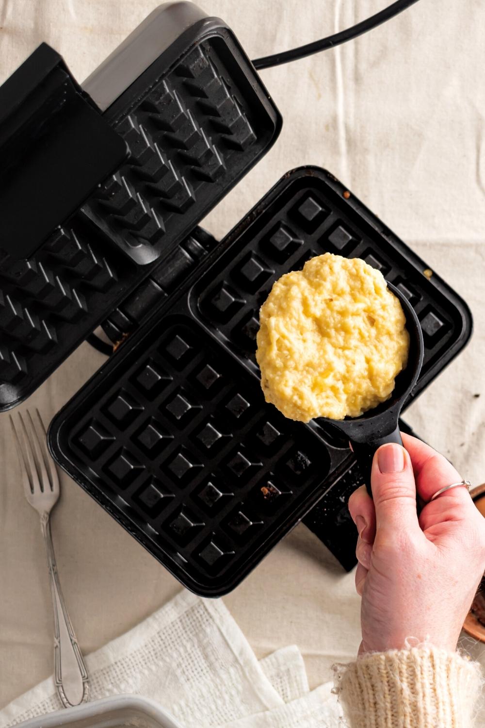 A hand holding a cup with banana waffle batter over a waffle iron.