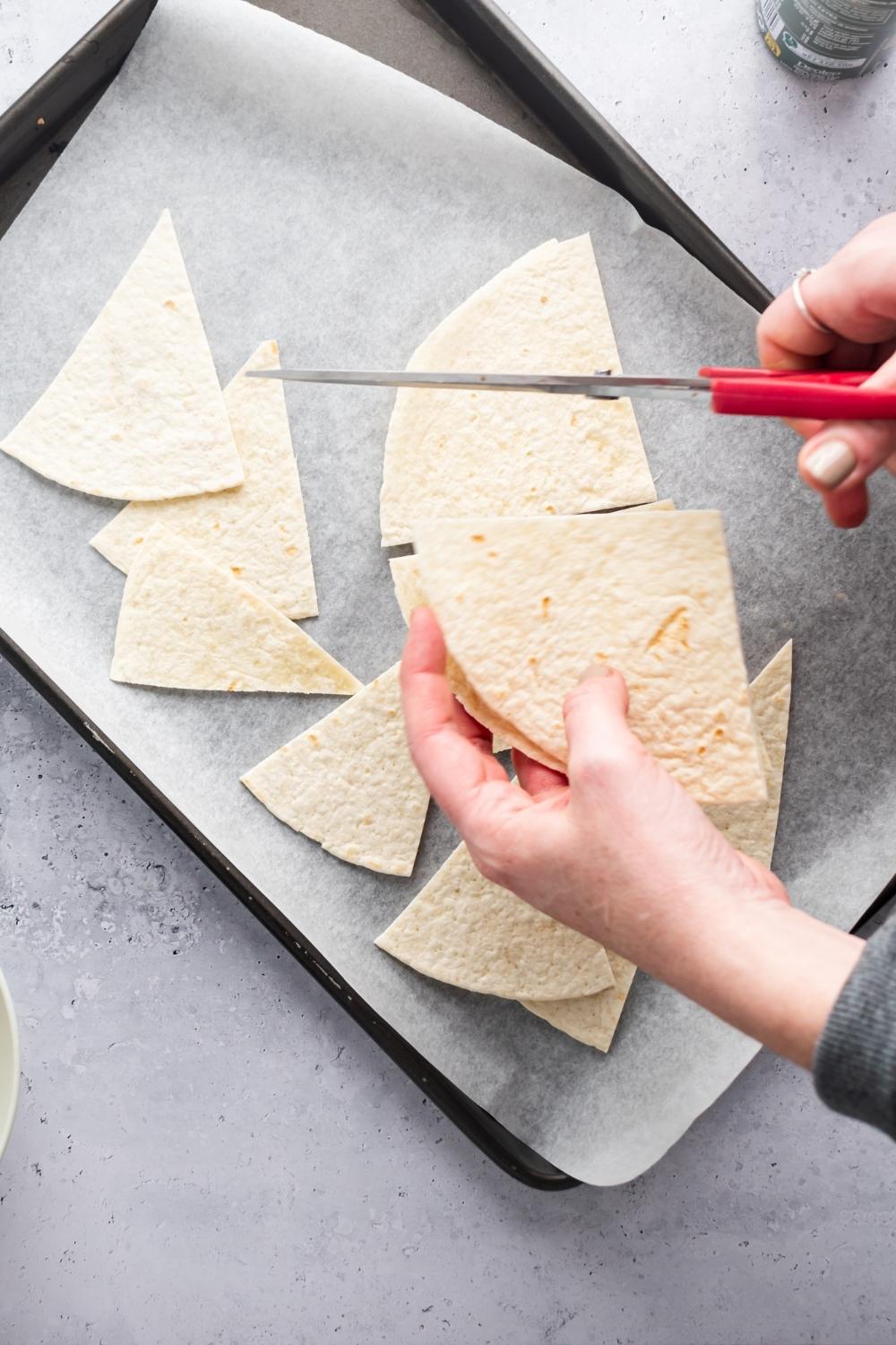 And hand holding a pair of scissors cutting a tortilla into triangles. There are a few triangular shaped pieces of tortilla below it on a piece of parchment paper on a baking sheet.