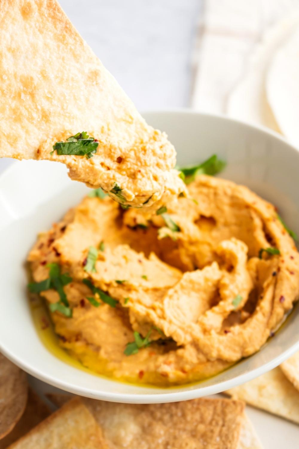 Part of a tortilla chip with hummus on the front of it hovering over spicy hummus in a white bowl.