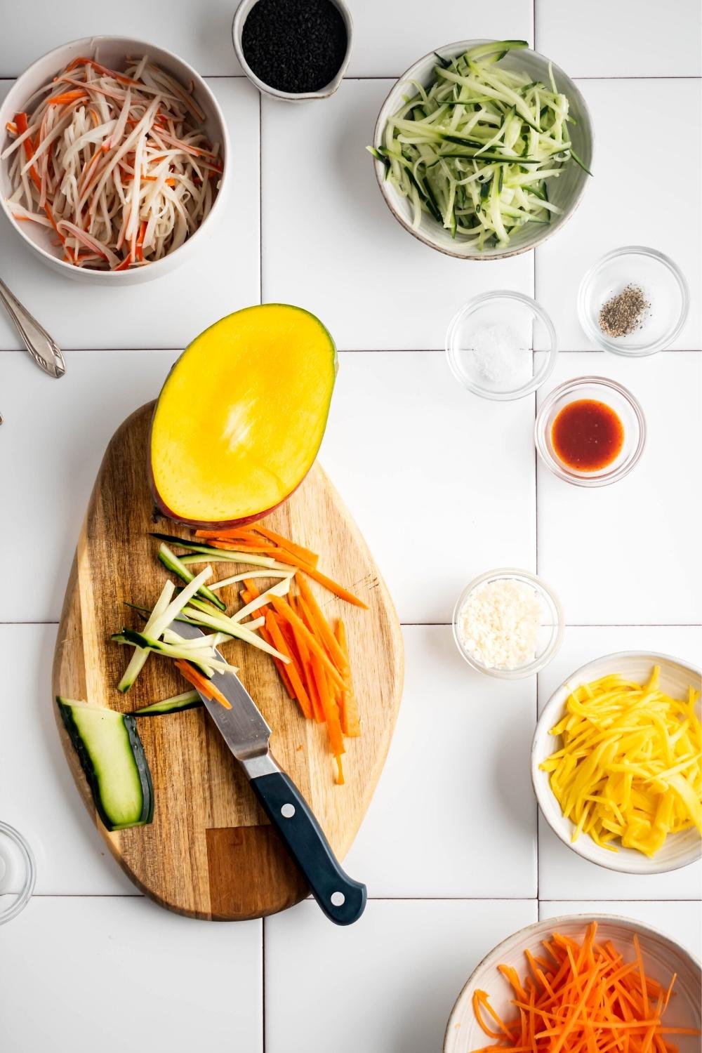 Some shredded cucumber, some shredded carrots, and half a mango on a wood cutting board on a white tile counter. Also on the counter is a bowl of imitation crab, a bowl of shredded cucumber, a small glass bowl of Sriracha, small glass bowl meal, a bowl shredded mango, and part of a bowl of shredded carrots.