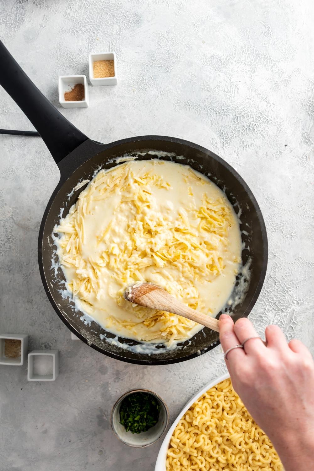 A hand holding a wooden spoon stirring around shredded cheese and sauce and a black skillet. In front of it is part of a white bowl that is filled with elbow macaroni noodles.