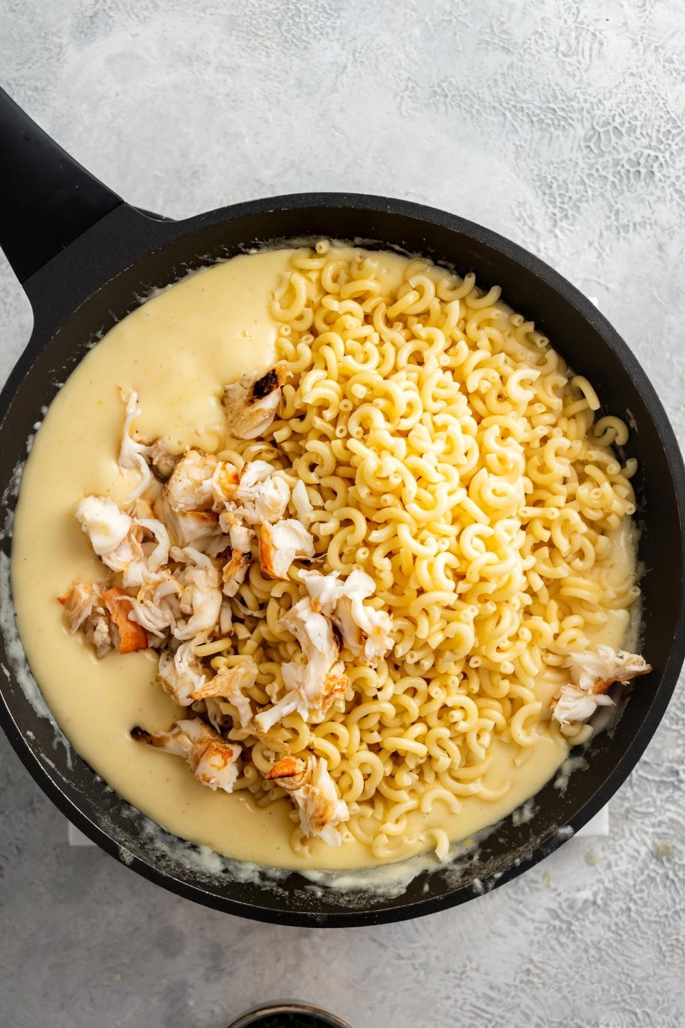 Elbow macaroni noodles, pieces of lobster, and cheese sauce all in a black skillet on a gray counter.