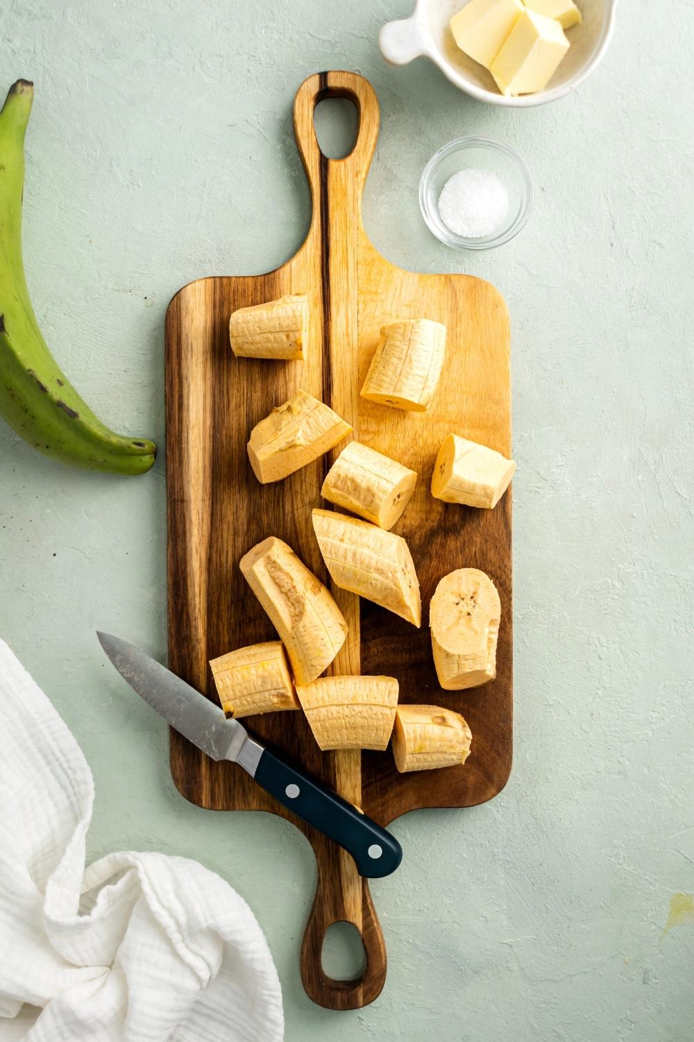 Slices of plantains and a knife on a wooden cutting board on top of the gray counter. Behind the cutting board is a small glass cup with sugar in it and part of a white bowl with cubed butter in it.