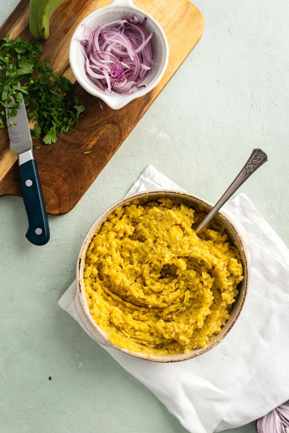 Mashed plantains in a bowl with a utensil submerged in it. The ball is on top of the white tablecloth on a gray counter behind it is chopped parsley and a white bowl of pickled red onion on a wooden cutting board.