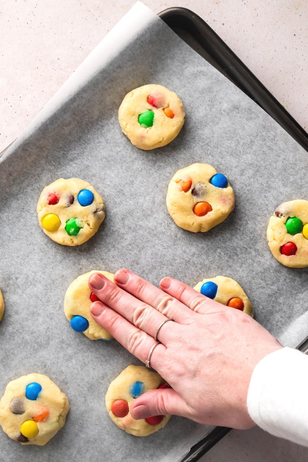 M&M sugar cookies on a piece of parchment paper on a baking tray. A hand is pressing the top of one of the cookies.