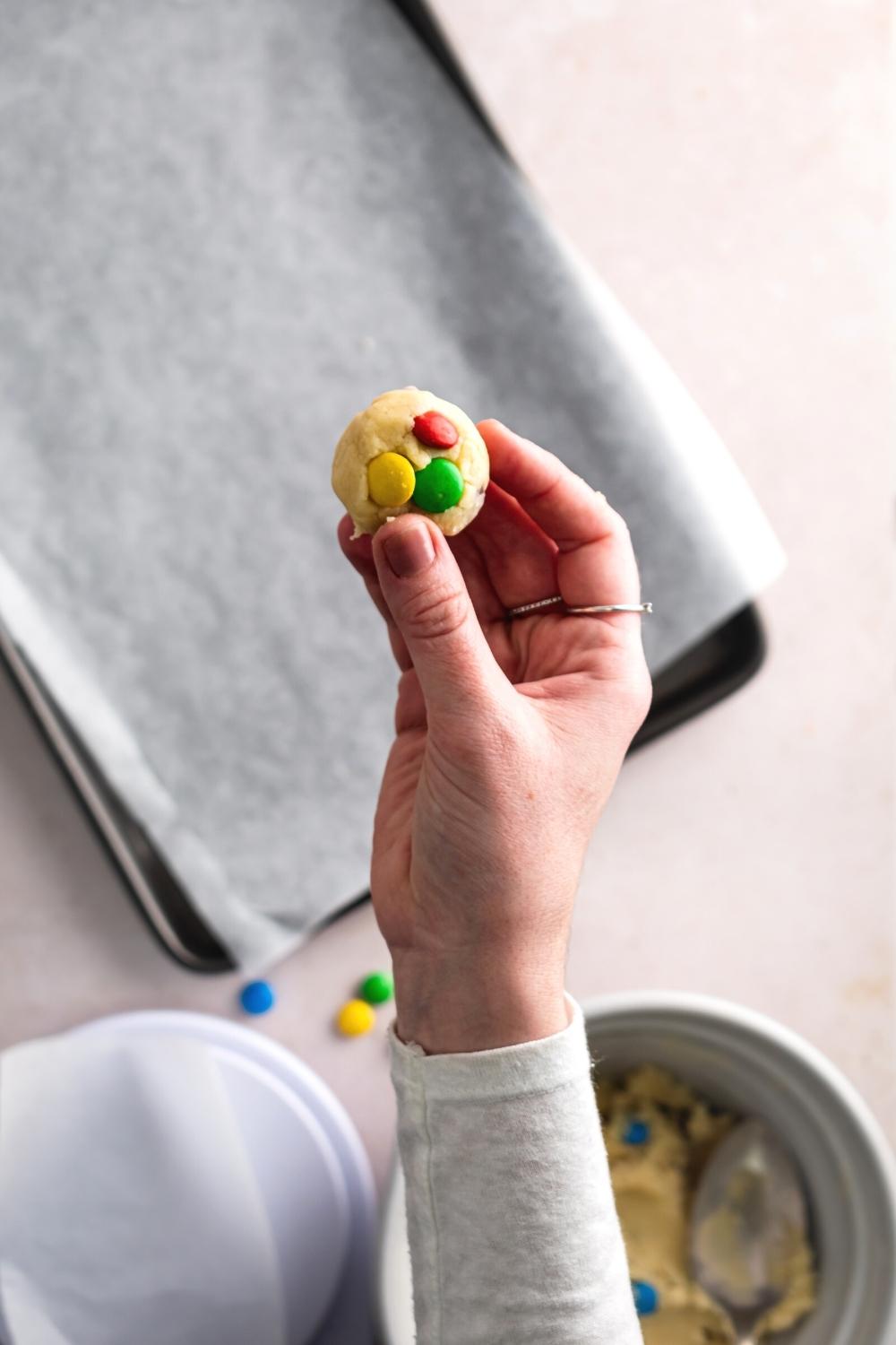 A hand holding a sugar cookie dough ball with three M&Ms in it.