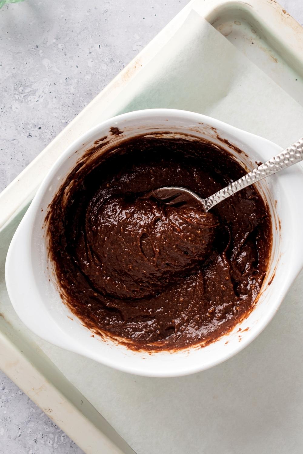 Nutella cookie dough with a spoon submerged in it in a white bowl on a piece of parchment paper on a baking sheet and a white counter.