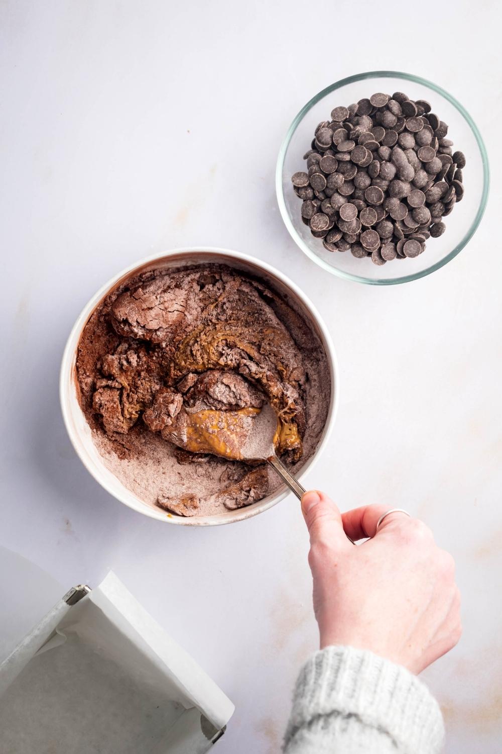 A hand holding a spoon mixing together protein brownie ingredients. Next to it is a small bowl of chocolate chips all on a white counter.