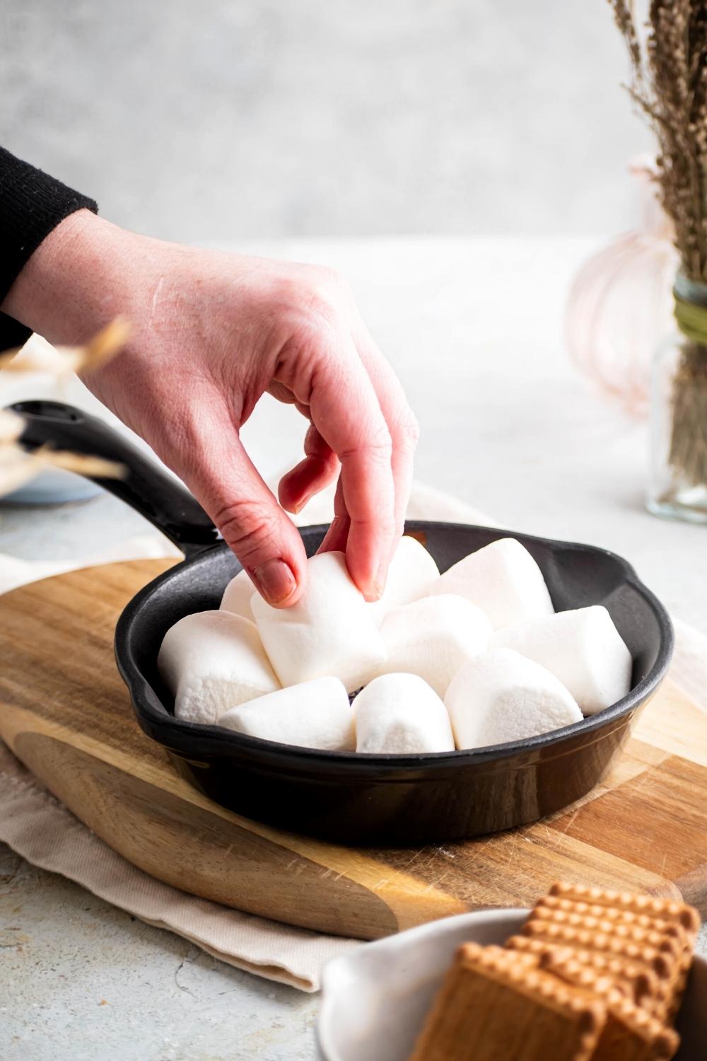 A hand holding a marshmallow that is touching a few other marshmallows all in a skillet on top of the wooden cutting board and a white tablecloth on a white counter.