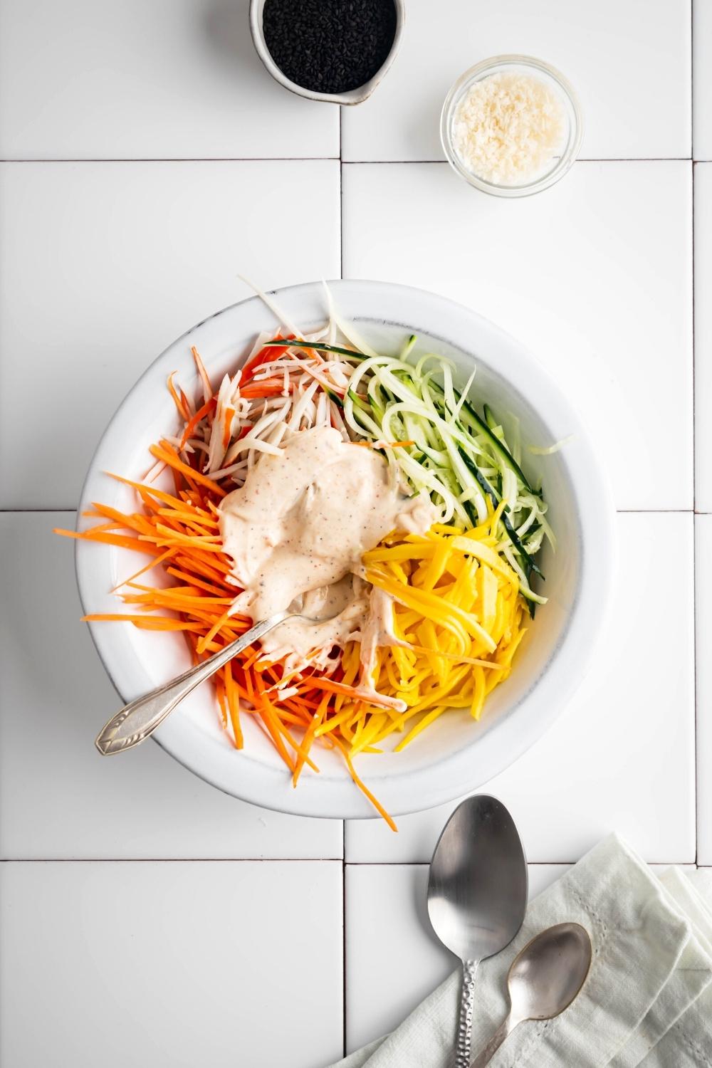 Kani salad dressing, julienned carrots, cucumber, mango, and imitation crab in a white bowl on a white tile counter.