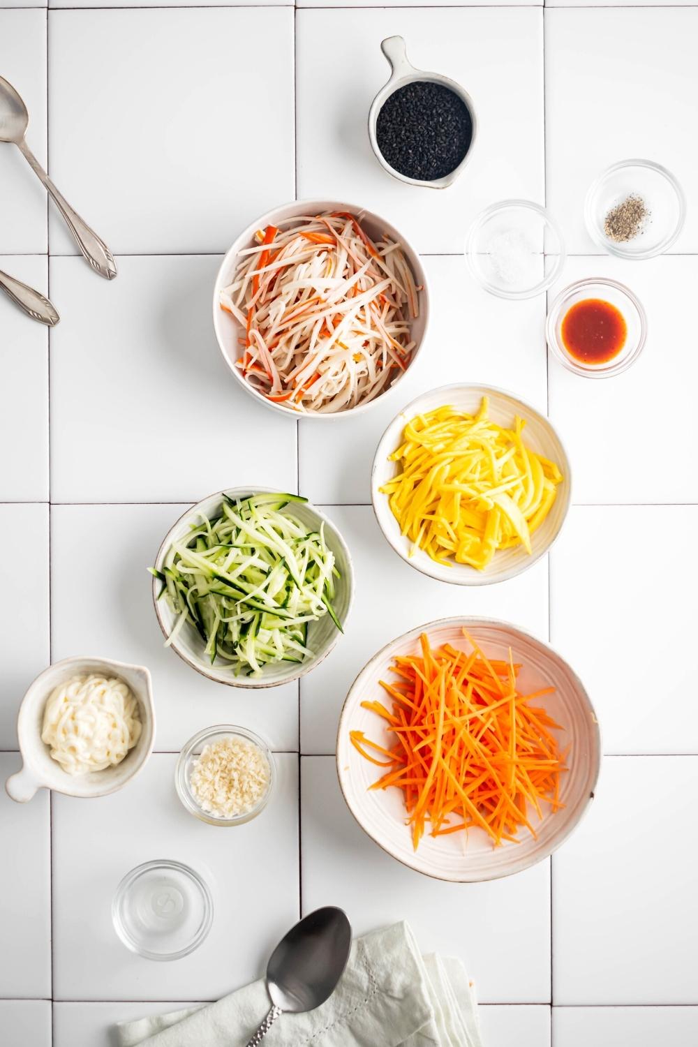 Shredded carrots in a white bowl, shredded cucumber in a white bowl, shredded mango in a white bowl, shredded imitation crab in a white bowl, a bowl of mayonnaise, a bowl of black sesame seeds, and a bowl of sriracha all on a white tile counter.