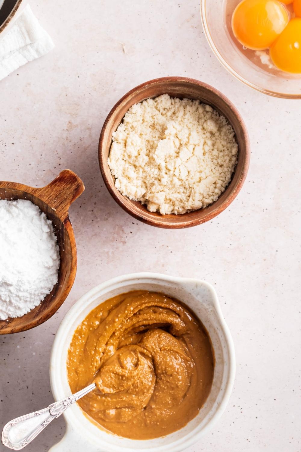 A bowl of peanut butter, a bowl of confectioner erythritol, a bowl of almond flour, and part of a bowl of four eggs all on a white counter.
