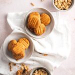 Two bowls of peanuts surrounding two bowls of keto peanut butter cookies on top of a tablecloth on a white counter.
